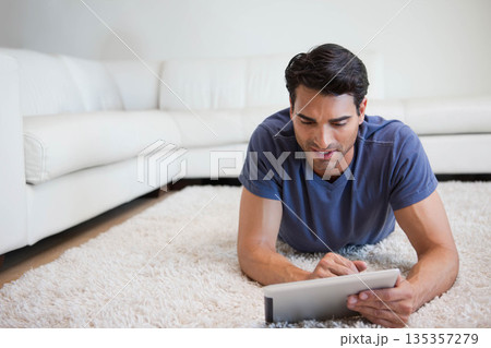 Mid-adult Hispanic male lying on white shag carpet at home holding tablet near white leather sofa 135357279