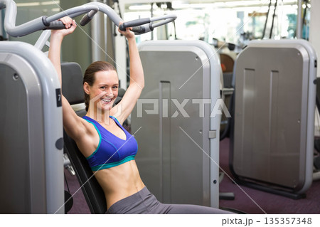 Adult woman wearing sports bra pushing chest press machine handles at gym by mirrors, copy space Adult woman wearing sports bra pushing chest press machine handles at gym by mirrors, copy space 135357348