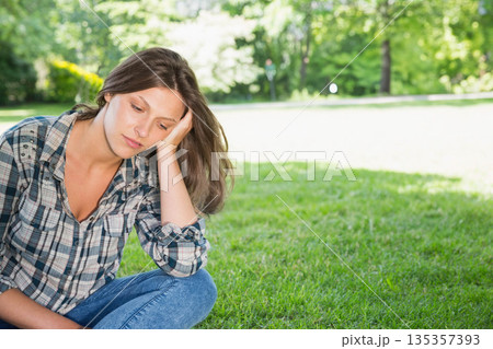 Teenage girl sitting cross-legged on grass in park, leaning head on hand under sunlight, copy space 135357393