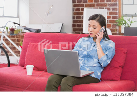 Chinese woman talking on smartphone while using laptop on sofa in loft with bicycle, copy space Chinese woman talking on smartphone while using laptop on sofa in loft with bicycle, copy space 135357394