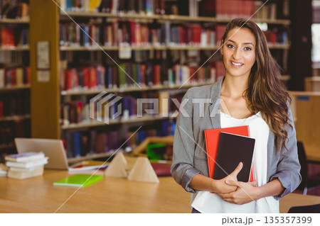 Female student standing by tables holding notebooks and using laptop in library area, copy space Female student standing by tables holding notebooks and using laptop in library area, copy space 135357399