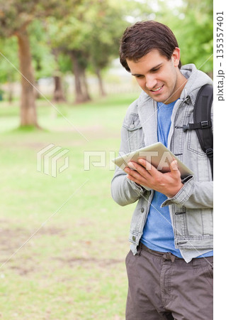 Man standing in park holding tablet and smiling while wearing jacket with backpack, copy space Man standing in park holding tablet and smiling while wearing jacket with backpack, copy space 135357401