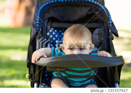 Infant boy gripping stroller tray bar and peeking over polka dot seat cushion in sunny park 135357427