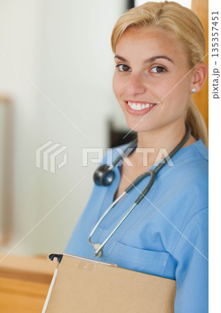 Female nurse in scrubs smiling while standing at nurse station, holding clipboard with stethoscope Female nurse in scrubs smiling while standing at nurse station, holding clipboard with stethoscope 135357451