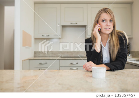 Female in blazer talking on smartphone near white mug while leaning on kitchen counter, copy space 135357457