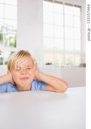 Boy pressing hands over ears and squinting at white tabletop inside room by grid-pattern window 135357464