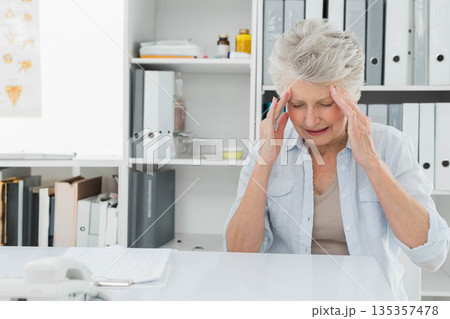 Senior woman pressing temples with hands at clinic desk with telephone and notebook, copy space Senior woman pressing temples with hands at clinic desk with telephone and notebook, copy space 135357478