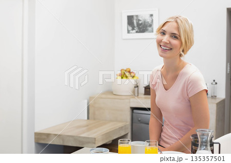 Blonde woman smiling while sitting at kitchen counter with orange juice glasses and coffee carafe Blonde woman smiling while sitting at kitchen counter with orange juice glasses and coffee carafe 135357501