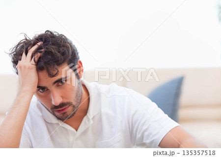Man sitting on beige couch by overexposed window in room running hand through hair with cushion Man sitting on beige couch by overexposed window in room running hand through hair with cushion 135357518