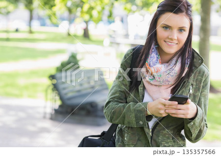 Woman wearing camouflage jacket, holding smartphone, carrying bag in sunlit park, copy space Woman wearing camouflage jacket, holding smartphone, carrying bag in sunlit park, copy space 135357566