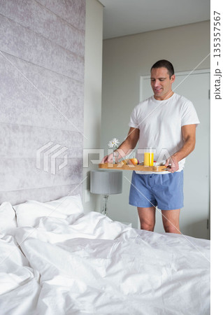 Man in thirties holding breakfast tray with orange juice, peach, daisy by unmade bed, copy space 135357567