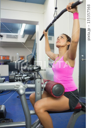 Woman gripping cable bar and pulling down lat pulldown machine at gym wearing pink wrist strap Woman gripping cable bar and pulling down lat pulldown machine at gym wearing pink wrist strap 135357568