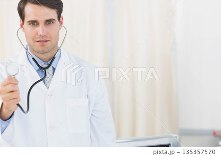 Male doctor wearing white coat holding stethoscope toward viewer in exam room with medical monitor 135357570