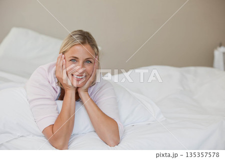 Smiling mid adult female lying on white bedding wearing light pink top hugging pillow in bedroom Smiling mid adult female lying on white bedding wearing light pink top hugging pillow in bedroom 135357578