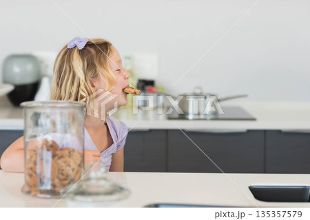 Child girl reaching for chocolate chip cookie from glass jar on white kitchen counter, copy space Child girl reaching for chocolate chip cookie from glass jar on white kitchen counter, copy space 135357579