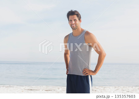 Man standing on sandy shore at water edge under blue sky, wearing gray tank, navy shorts 135357603