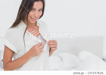 Woman holding white button-up shirt revealing lace-trimmed camisole in bright bedroom, copy space 135357626