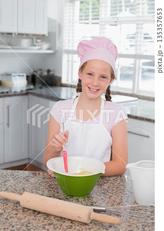 Child girl wearing pink chef hat and apron stirring batter in mixing bowl in residential kitchen 135357653