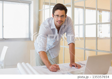 Male professional leaning over table in office drawing blueprints with pencil next to open laptop 135357654
