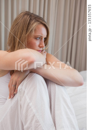 White woman sitting hugging knees on bed in bedroom with bedding beige curtains left-hand ring 135357655