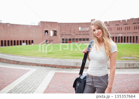 Female student posing with messenger bag beside concrete border with brick facade, copy space 135357664