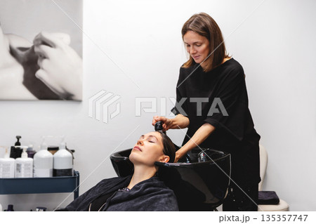 Professional hairstylist washing hair of woman in modern beauty salon, soothing cleansing treatment in white interior Professional hairstylist washing hair of woman in modern beauty salon, soothing cleansing treatment in white interior 135357747