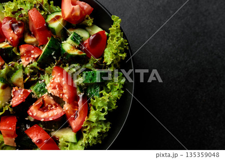 Vegetable salad of tomatoes, cucumbers, onions, lettuce dressed with aromatic dressing in a black plate on a gray background Vegetable salad of tomatoes, cucumbers, onions, lettuce dressed with aromatic dressing in a black plate on a gray background 135359048