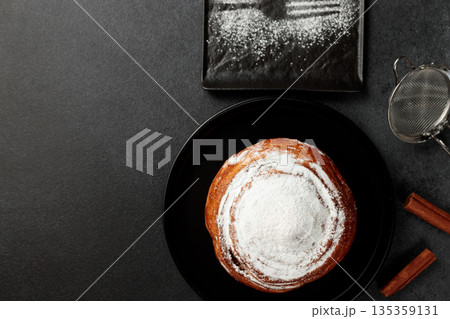 Round, delicious bun with cinnamon and powdered sugar on a black plate on a gray background Round, delicious bun with cinnamon and powdered sugar on a black plate on a gray background 135359131