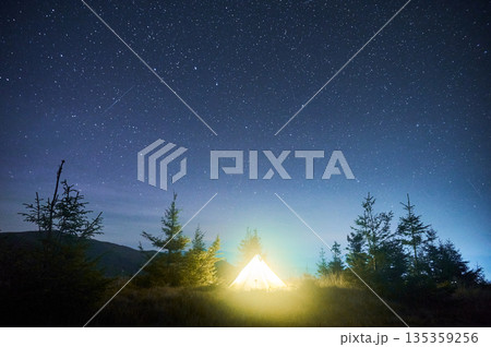 Glowing yellow tent under sky filled with countless stars, including visible shooting star. Surrounded by pine trees on grassy hill, serene scene captures magic of peaceful night in nature. 135359256