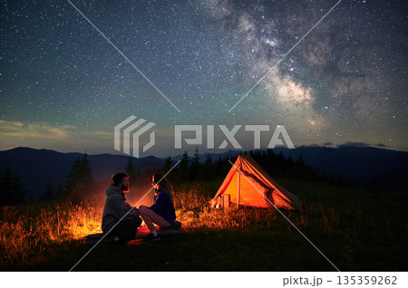 Couple having rest by campfire near their illuminated tent beneath stunning star-filled sky. Milky Way stretches across heavens, creating magical and serene ambiance in mountainous wilderness. 135359262