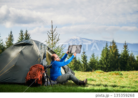 Man sits beside tourist tent, holding portable solar panel charger to capture sunlight for charging power bank. Large orange backpack, camera on tripod. Snow-capped mountains under partly cloudy sky. Man sits beside tourist tent, holding portable solar panel charger to capture sunlight for charging power bank. Large orange backpack, camera on tripod. Snow-capped mountains under partly cloudy sky. 135359268