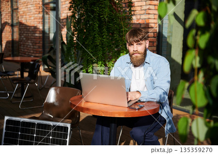 Man works on laptop and smartphone at outdoor table. Solar panel nearby, emphasizing sustainable, modern eco-friendly workspace that combines technology and sustainability. Man works on laptop and smartphone at outdoor table. Solar panel nearby, emphasizing sustainable, modern eco-friendly workspace that combines technology and sustainability. 135359270