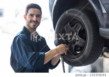 Male mechanic standing by raised car wheel removing lug nuts with pneumatic wrench in garage 135359291