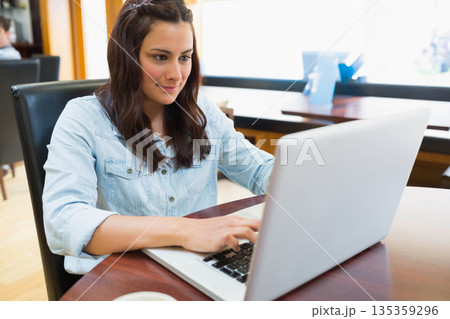 Woman typing on silver laptop at cafe, sitting beside white coffee cup on saucer Woman typing on silver laptop at cafe, sitting beside white coffee cup on saucer 135359296