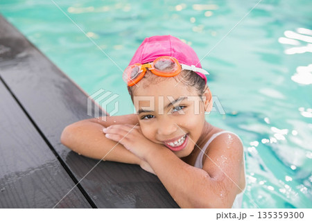 African American girl leaning on pool edge over turquoise water smiling in pink cap, orange goggles 135359300