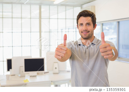 Man giving two thumbs-up standing in bright modern office with desks, monitors and glass partition Man giving two thumbs-up standing in bright modern office with desks, monitors and glass partition 135359301