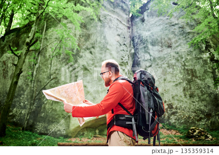 Bearded tourist man studies map while stands in front of large rock in dense forest. Traveler with grey backpack and glasses, planning outdoor adventure, hiking or climbing route. 135359314