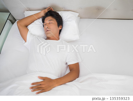 Japanese man sleeping in bedroom, resting head on pillow with duvet beside table, copy space Japanese man sleeping in bedroom, resting head on pillow with duvet beside table, copy space 135359323