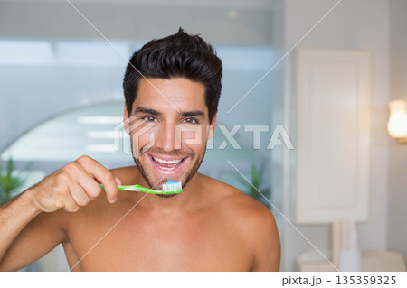 Shirtless adult man smiling while brushing teeth at bathroom sink with green toothbrush 135359325
