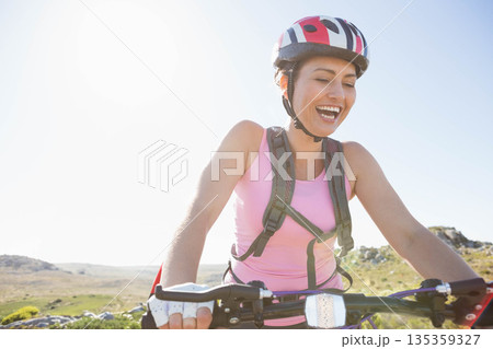 Mid 30s woman pedaling on trail holding handlebars wearing hydration pack, cycling helmet, gloves Mid 30s woman pedaling on trail holding handlebars wearing hydration pack, cycling helmet, gloves 135359327