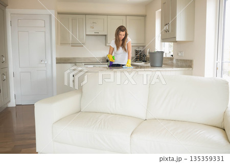 Female wearing yellow gloves wiping granite countertop with sponge and cloth in kitchen area 135359331