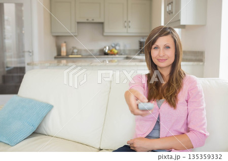 Woman holding remote control sitting on sofa with pillow by kitchen counter fruit bowl, copy space Woman holding remote control sitting on sofa with pillow by kitchen counter fruit bowl, copy space 135359332