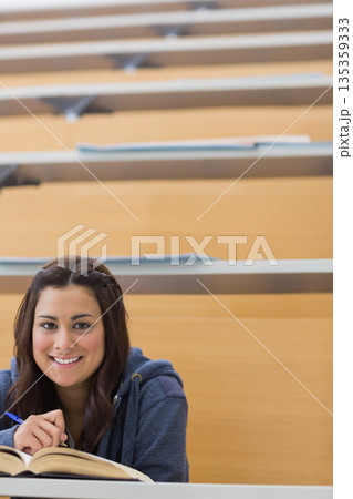 Female student sitting at lecture hall desk holding blue pen at open textbook, copy space Female student sitting at lecture hall desk holding blue pen at open textbook, copy space 135359333
