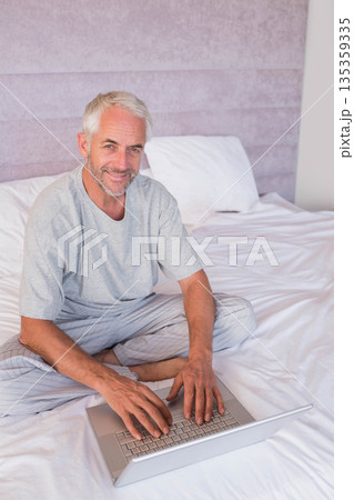 Senior man sitting cross-legged on white linens at home using silver laptop against grey headboard Senior man sitting cross-legged on white linens at home using silver laptop against grey headboard 135359335