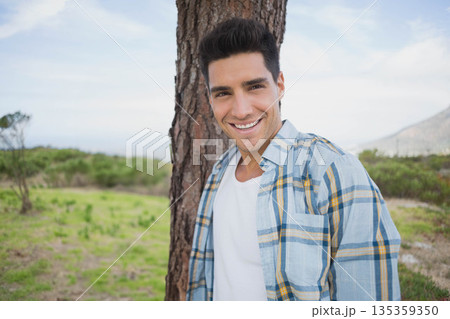 Man in his twenties leaning against tree trunk in grassy clearing with rolling hills 135359350
