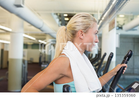 Woman in turquoise tank top wearing gym towel exercising on elliptical machine by ducts, copy space Woman in turquoise tank top wearing gym towel exercising on elliptical machine by ducts, copy space 135359360