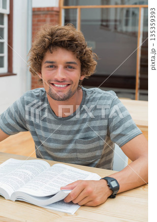 Male student sitting at table in library holding open reference book wearing digital wristwatch Male student sitting at table in library holding open reference book wearing digital wristwatch 135359393
