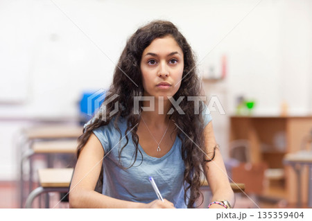 Female student writing notes while listening to instructor in classroom with pen and notebook 135359404