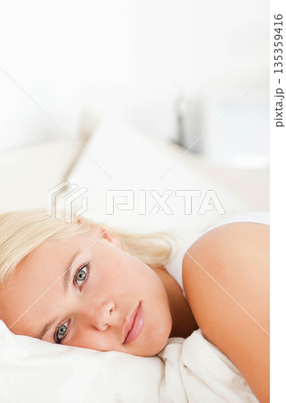 Woman lying on bed in bright bedroom wearing white tank top with white pillow and sheets 135359416