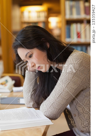Asian woman leaning on hand reading open textbook at wooden library study table with notebook pen Asian woman leaning on hand reading open textbook at wooden library study table with notebook pen 135359427